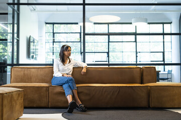 Indian woman sitting on sofa with ottoman in office lounge by glass partition under pendant lights