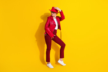 Confident mature woman in red leather jacket and hat posing stylishly against vibrant yellow background