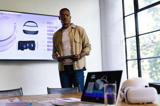 African American man presenting VR headset models on tablet at conference table with wall display
