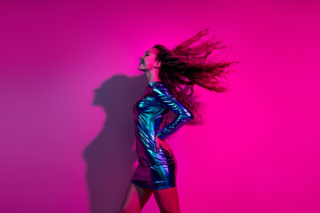 Dynamic woman in glossy blue dress smiling against vibrant pink neon background celebrating nightlife
