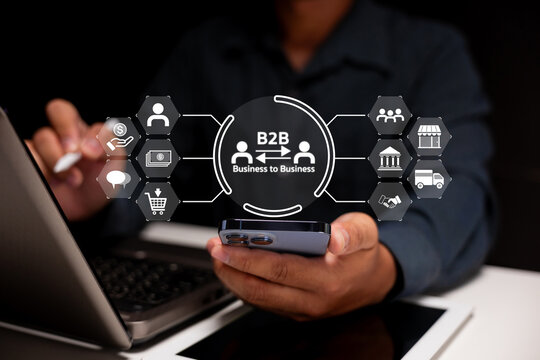 A person uses a smartphone and laptop with a digital B2B business-to-business concept interface displaying various business and logistics icons. - Powered by Adobe