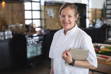Female chef in chef coat holding tablet beside vegetables on cutting board in restaurant kitchen