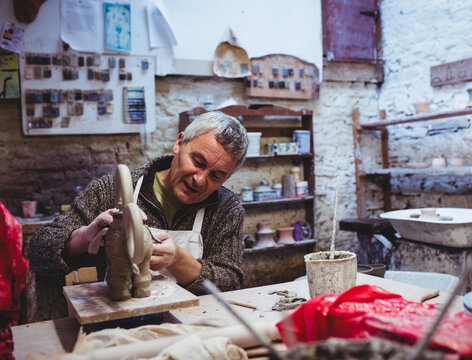Middle-aged man shaping clay sculpture at rustic pottery studio with sculpting tools and glaze jars