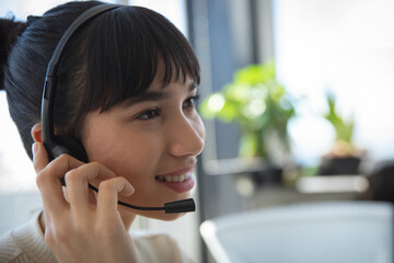 Female support representative smiling and holding headset at desk by window with plants, copy space