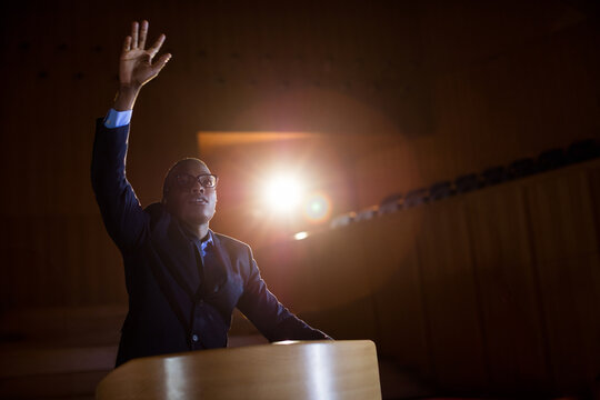 African American man standing behind podium on stage raising right hand under spotlight, copy space
