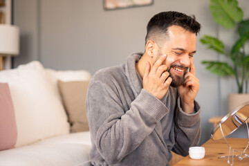 Cheerful Mid-Aged Man Wearing Comfortable Grey Robe Applying Moisturizer During Morning Self-Care...