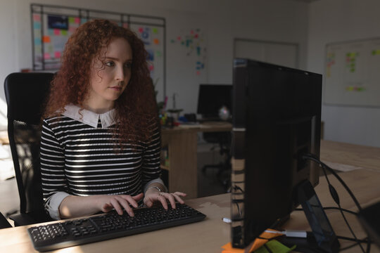 Woman with curly hair typing on full-size keyboard at office desk with whiteboard and sticky notes