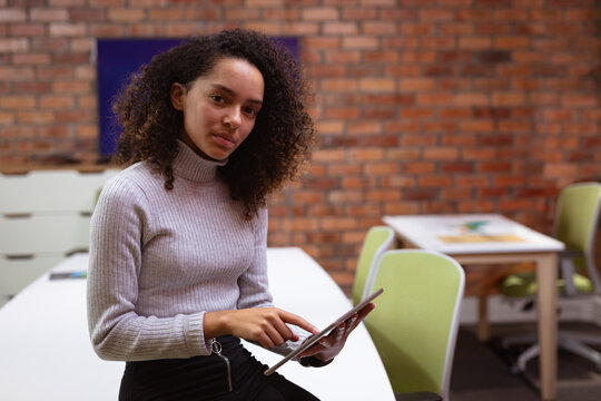 African American woman using tablet on white desk in office with green chairs, exposed brick wall
