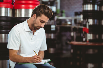 Man writing on clipboard with pencil among kegs with red lids in brewery warehouse, copy space