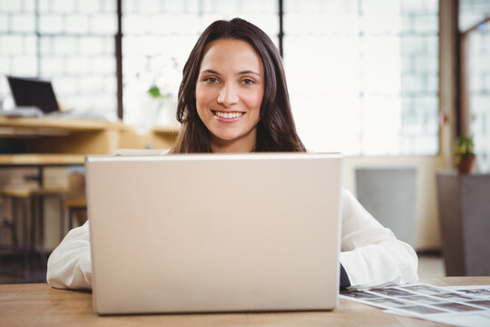 Woman sitting at wooden desk in office working on laptop with printed documents by grid windows