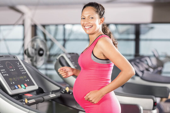 Pregnant woman walking on treadmill in modern gym wearing pink activewear with digital console