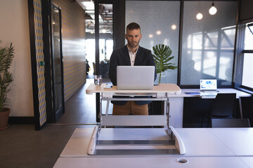 Mid adult man in business attire standing at adjustable desk typing on silver laptop in office