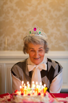 Senior woman wearing tiara blowing out lit candles on birthday cake at dining table