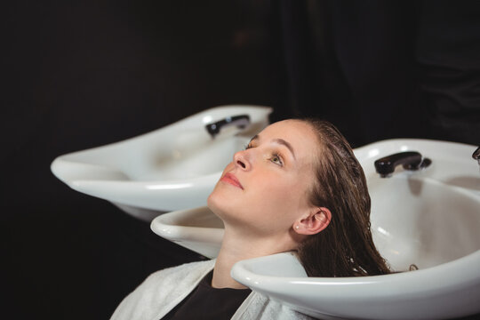 Female client reclining in salon shampoo chair with stylist rinsing hair in white ceramic basin