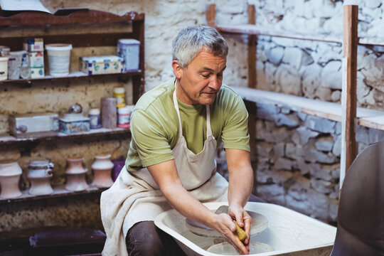 Male potter kneeling at pottery wheel in rustic stone workshop shaping clay with sponge - Powered by Adobe