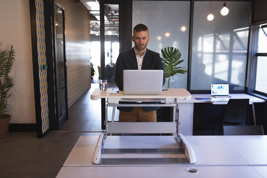 Mid adult man in business attire standing at adjustable desk typing on silver laptop in office