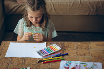 Female child holding yellow crayon while drawing on white paper at living room table with pastels
