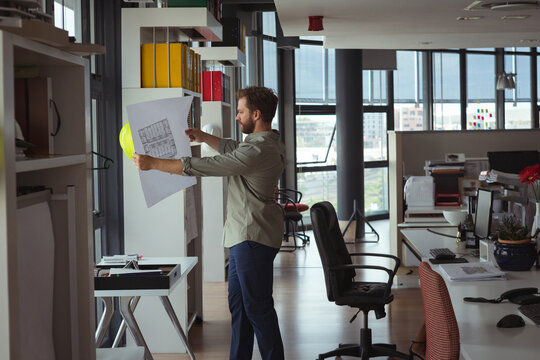 Man examining architectural blueprint in open office, showing shelving binders helmet, copy space