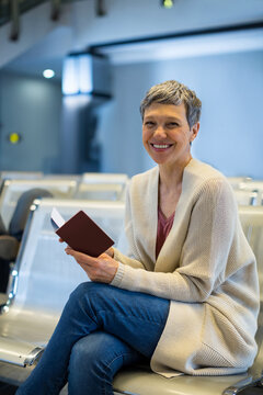 Mature woman holding open passport sitting on bench by flight display in airport lounge, copy space