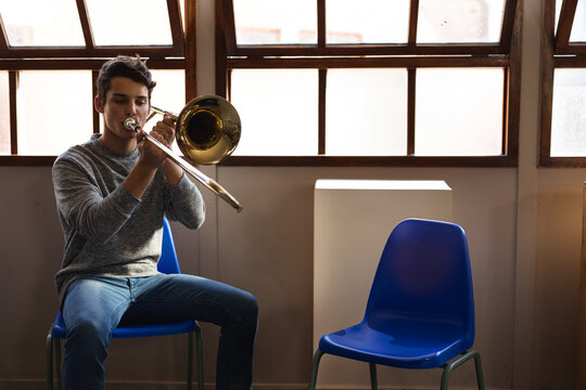 Man in twenties sitting on blue chairs playing trombone with pedestal in practice room, copy space