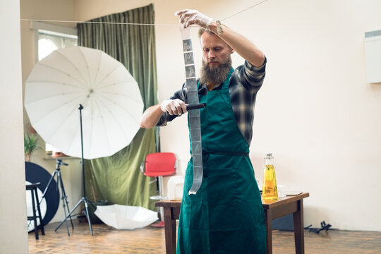 Male technician wearing green apron trimming film negatives on wire line in darkroom studio