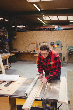 Mature male carpenter wearing red suspenders measuring long wooden plank on table saw in workshop