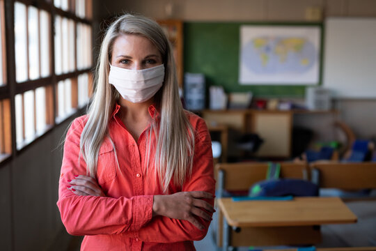 Teacher standing with arms crossed wearing face mask in classroom with desks windows and world map