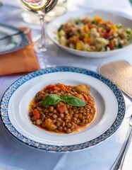 Fototapeta premium Delicious lentil stew served on a patterned plate with a fresh basil garnish, alongside a vibrant salad and wine glass on a white tablecloth, perfect for lunch or dinner.