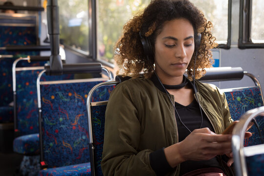 African American woman sitting on patterned bus seat wearing headphones scrolling smartphone