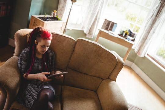 Woman sitting on brown sofa in sunlit living room corner using tablet wearing earphones, copy space