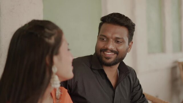An affectionate Indian couple shares an intimate moment outdoors, dressed in traditional ethnic attire. The woman wears a bright orange salwar suit while the man is in a black shirt and jeans. 