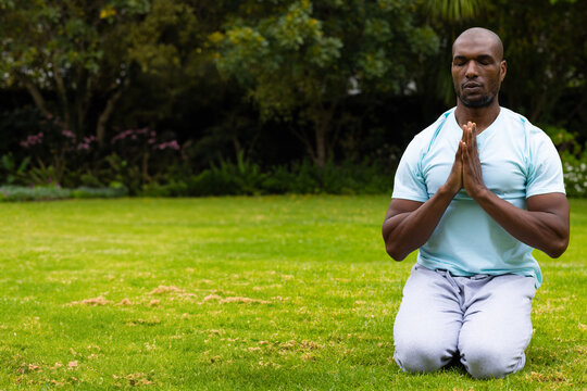 African American man kneeling on lawn pressing hands together in meditation at park, copy space