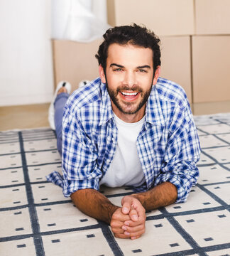 Man smiling and reclining on patterned rug in empty room with stacked cardboard boxes and sneakers