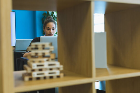 African American woman typing on laptop in office at desk amid wooden shelving and blocks