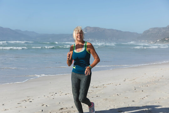 Senior woman jogging on sandy beach wearing neon trimmed tank top, leggings and running shoes