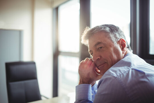 Man wearing blue dress shirt leaning at office desk next to chair and windows, copy space