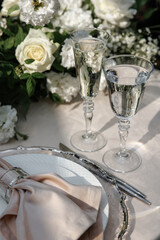 Elegant table setting with two glass goblets surrounded by white floral arrangements. Delicate details include a silver cutlery set and soft-colored napkin, ideal for weddings or celebrations.