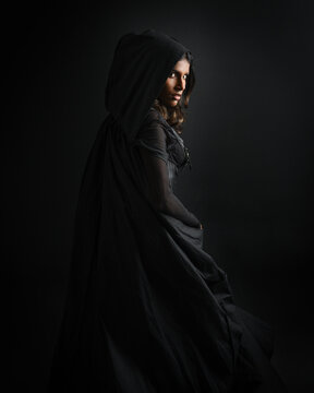  portrait of mysterious hooded woman in black cloak,  gothic fantasy or dystopian character. posing with gestural magical hand poses.  lit with moody shadows, studio scene on dark isolated background