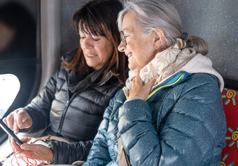 Cheerful modern senior couple of women sitting on a bus while looking together at smartphone sharing tech and social