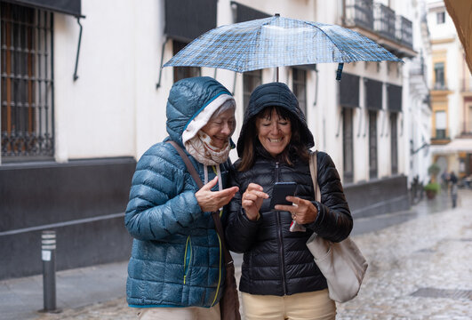 Joyful friendship in the rain. Two elderly women taking sightseeing tour and checking the right direction on their cell phones, standing under an umbrella