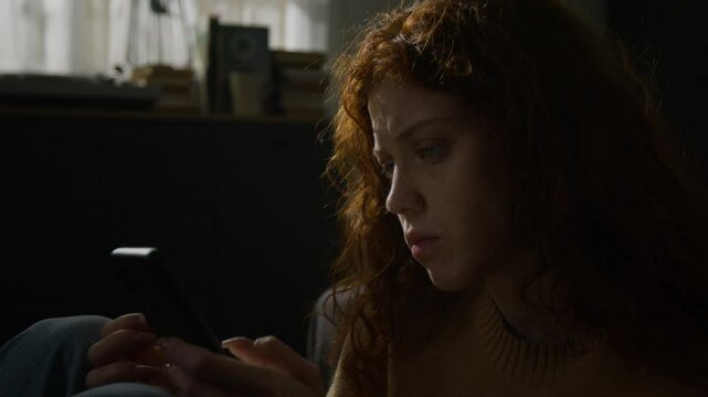 Stressed red-haired woman biting her nails and texting on her smartphone, while sitting on bedroom floor