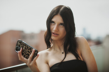 A stylish woman holds a phone while sitting on a balcony. She gazes thoughtfully into the distance, surrounded by a vibrant city backdrop under clear skies