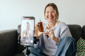 Middle-aged woman beauty blogger filming skincare product unboxing at home with ring light....