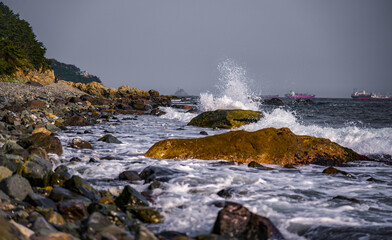 Ocean wave crashing on rocky shore at sunset splashes in motion with distant ships on horizon. Golden hour seascape dynamic wave action on stone shore with ships sailing into sunset. 