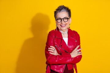 Confident mature woman in vibrant red leather jacket posing against a bright yellow backdrop with a charming smile