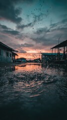 Fototapeta premium Sunset reflected in a rain-filled puddle, small buildings flank the scene, water droplets suspended in the air