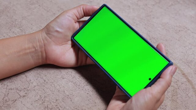 Hands holding a smartphone with green screen in horizontal position, ideal for adding custom content or mockups, Shot on a neutral surface for tech and advertising use, close-up