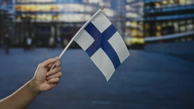 Hand holding finland flag in city street scene with blurred urban background during dusk representing national pride and culture.