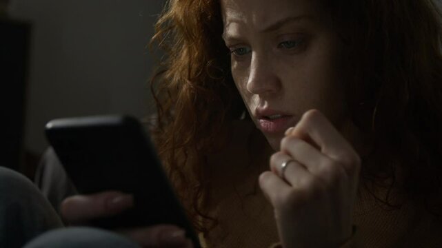 Close-up shot of anxious red-haired woman looking at smartphone screen while sitting on bedroom floor