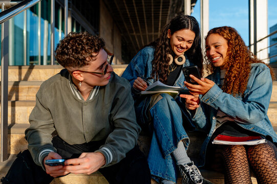 Three cheerful university students are sitting on campus stairs, sharing funny social media content on a smartphone and enjoying their time together